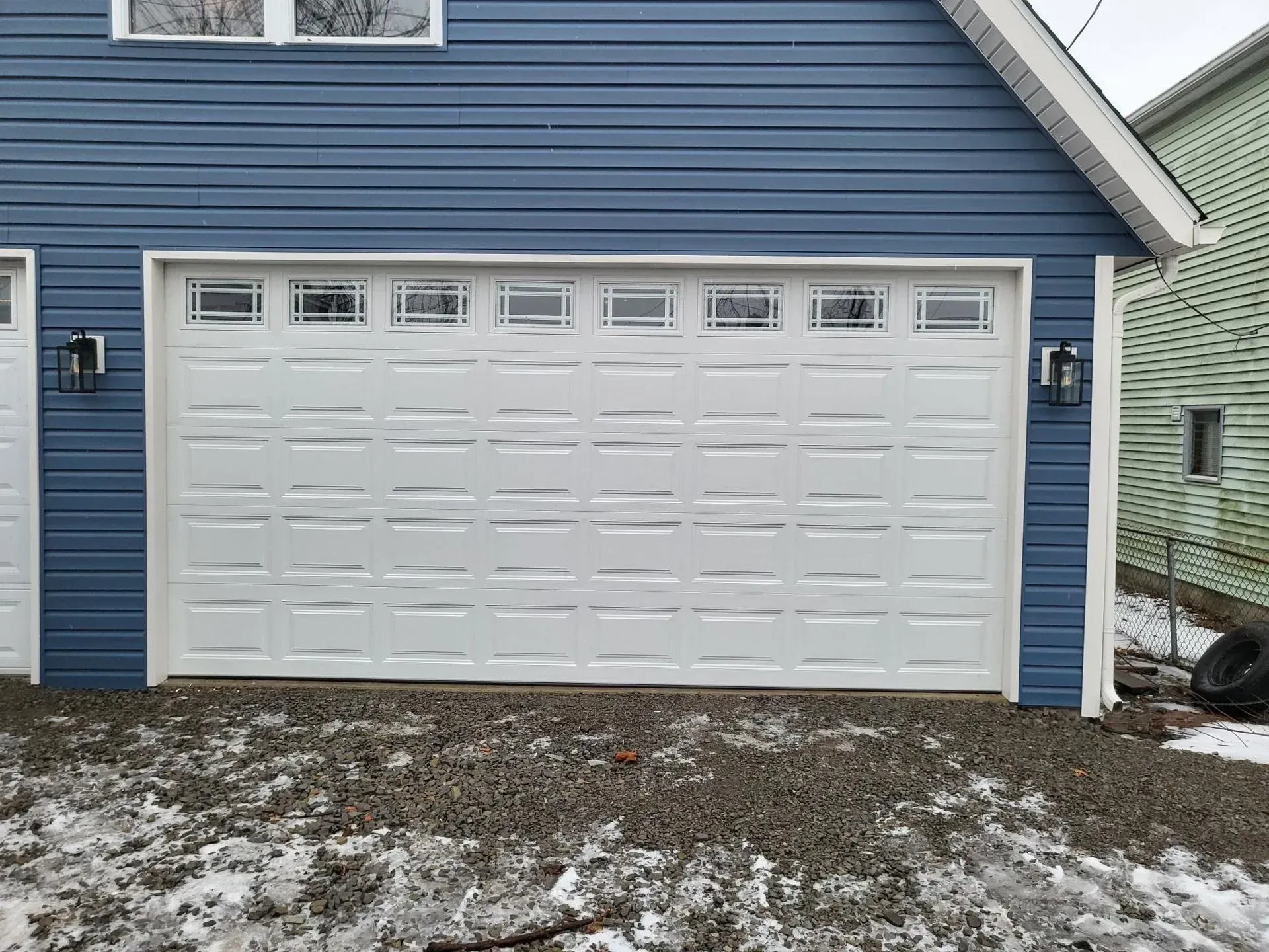White garage door with glass panes above, blue siding, gravel ground.