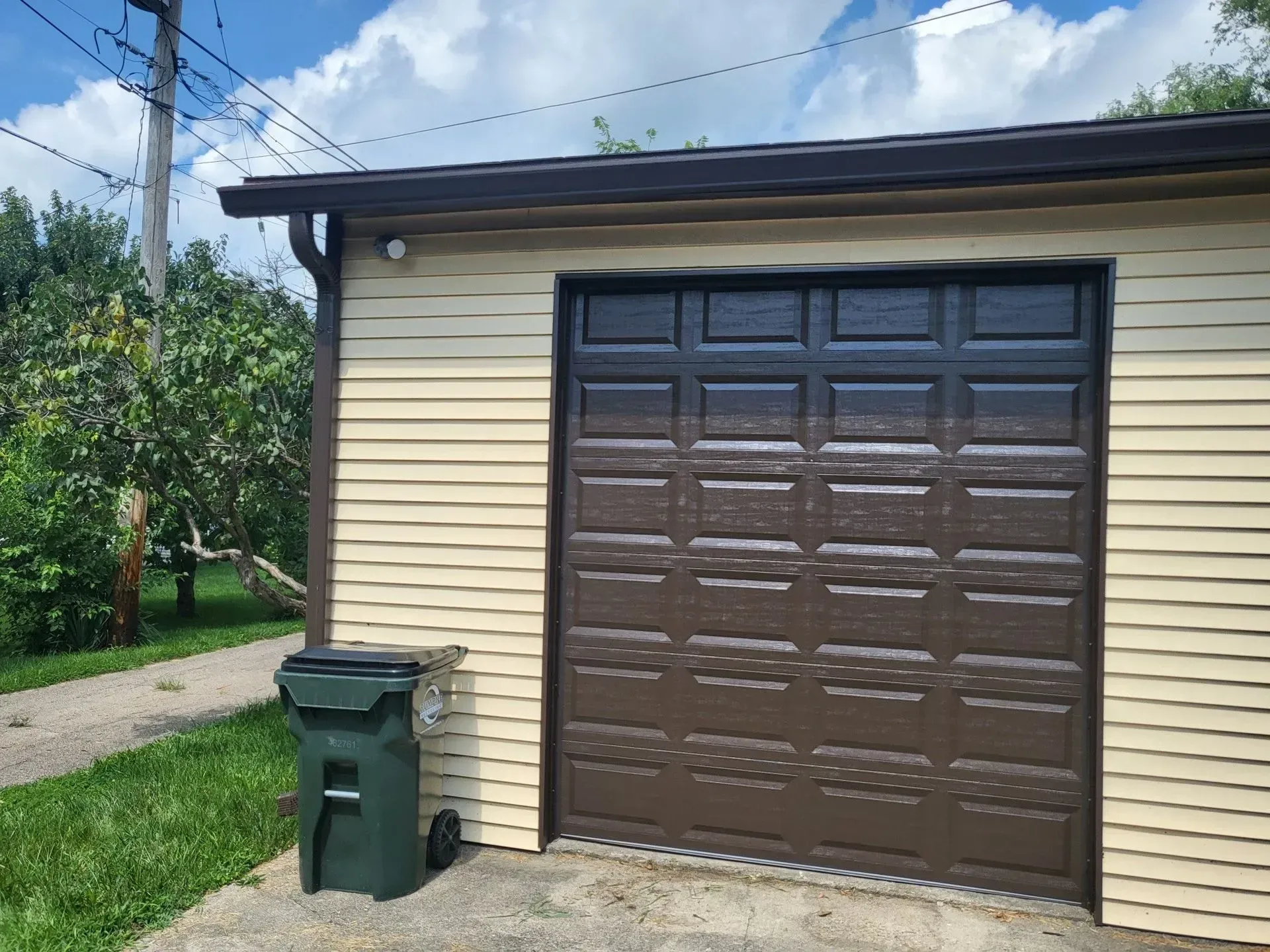 Beige garage with brown door and trim, green trash can, blue sky.