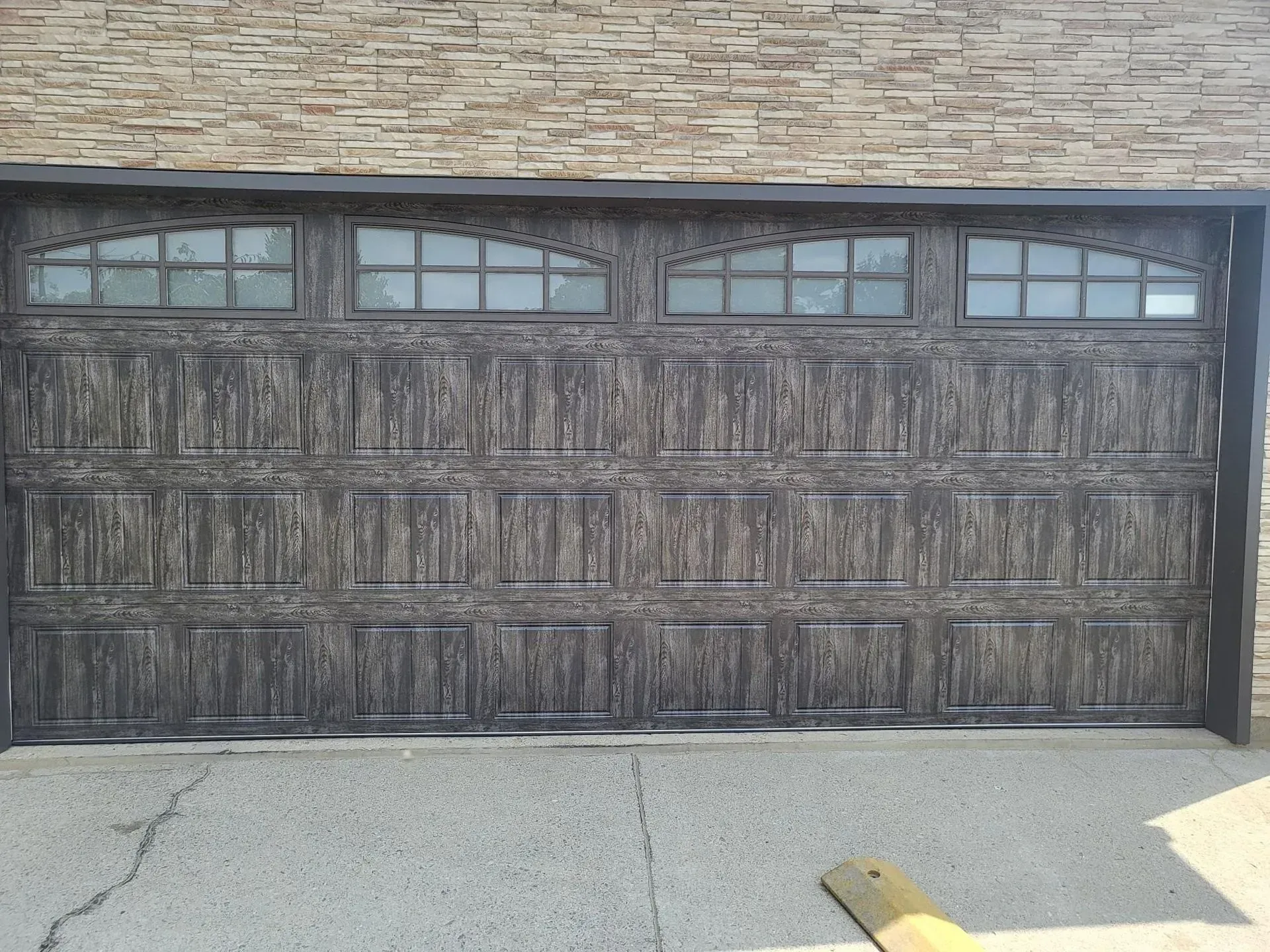 Dark gray garage door with arched windows and textured panels, set in a brick wall.