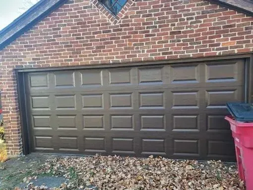 Brown garage door on a brick building with fallen leaves and a red trash can.