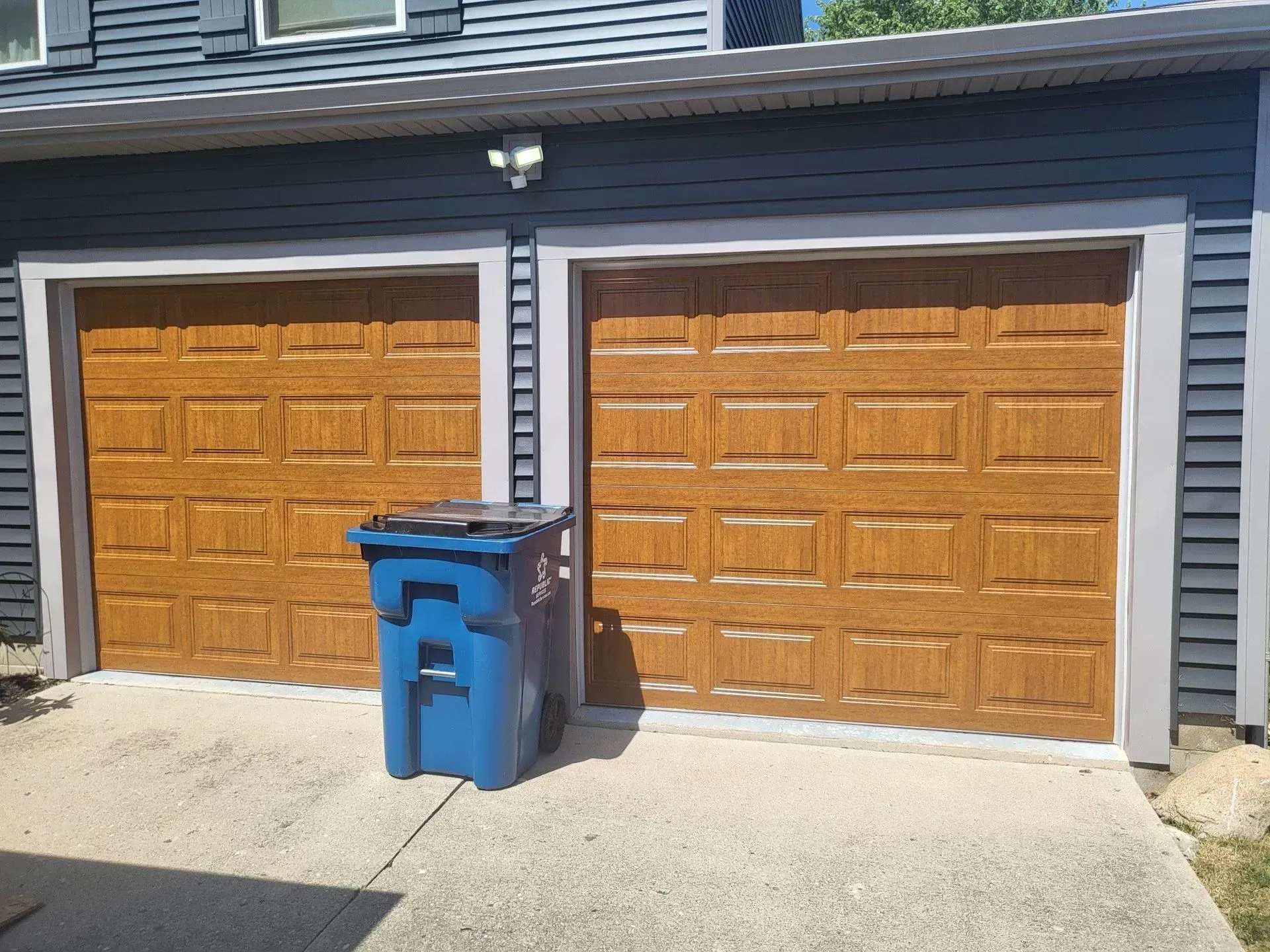 Two brown garage doors, blue trash bin on concrete driveway, blue siding, gray trim.