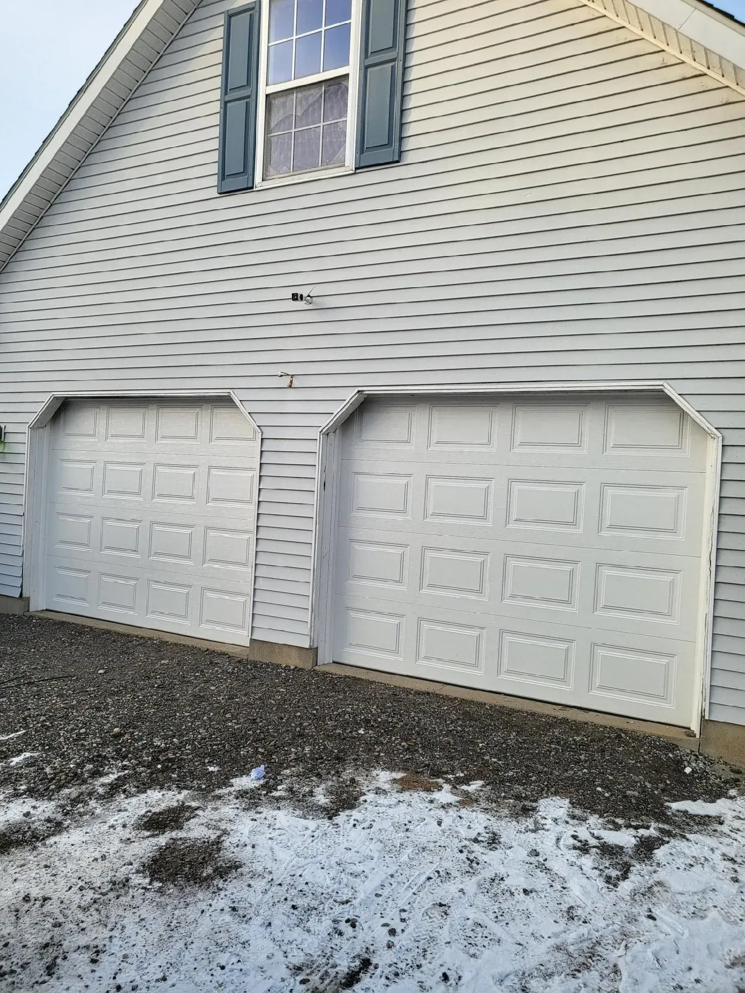 Two white garage doors on a light gray house with a window above. Snow covers the ground.