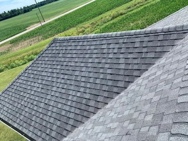 Gray shingled roof of a house with a green field in the background.