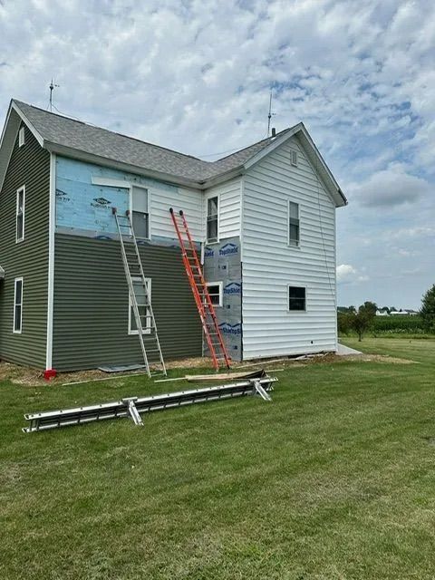 House exterior with green and white siding being renovated; ladders and materials on lawn.