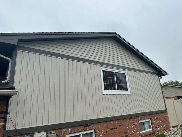 Side of a house with light tan siding, a dark gray roof, and a white-framed window on a cloudy day.
