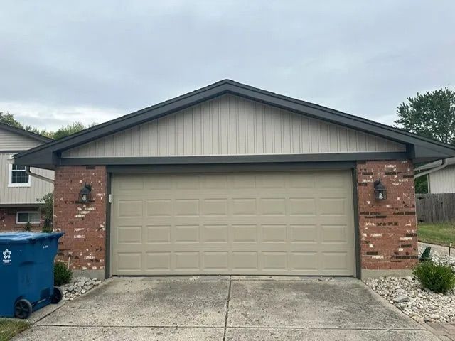 Garage with tan door, brick columns, and gray roof under a cloudy sky.