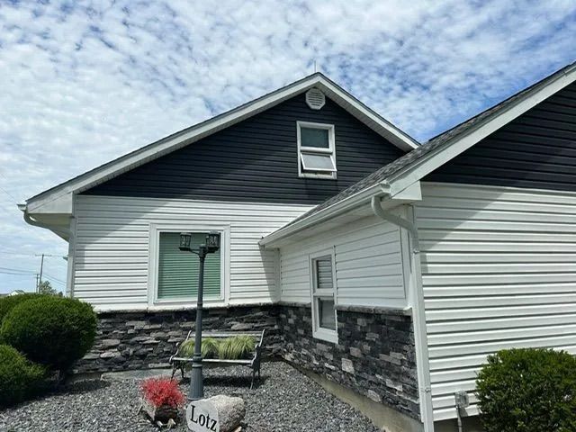 Two-story house with black and white siding, stone base, and green landscaping under a cloudy sky.