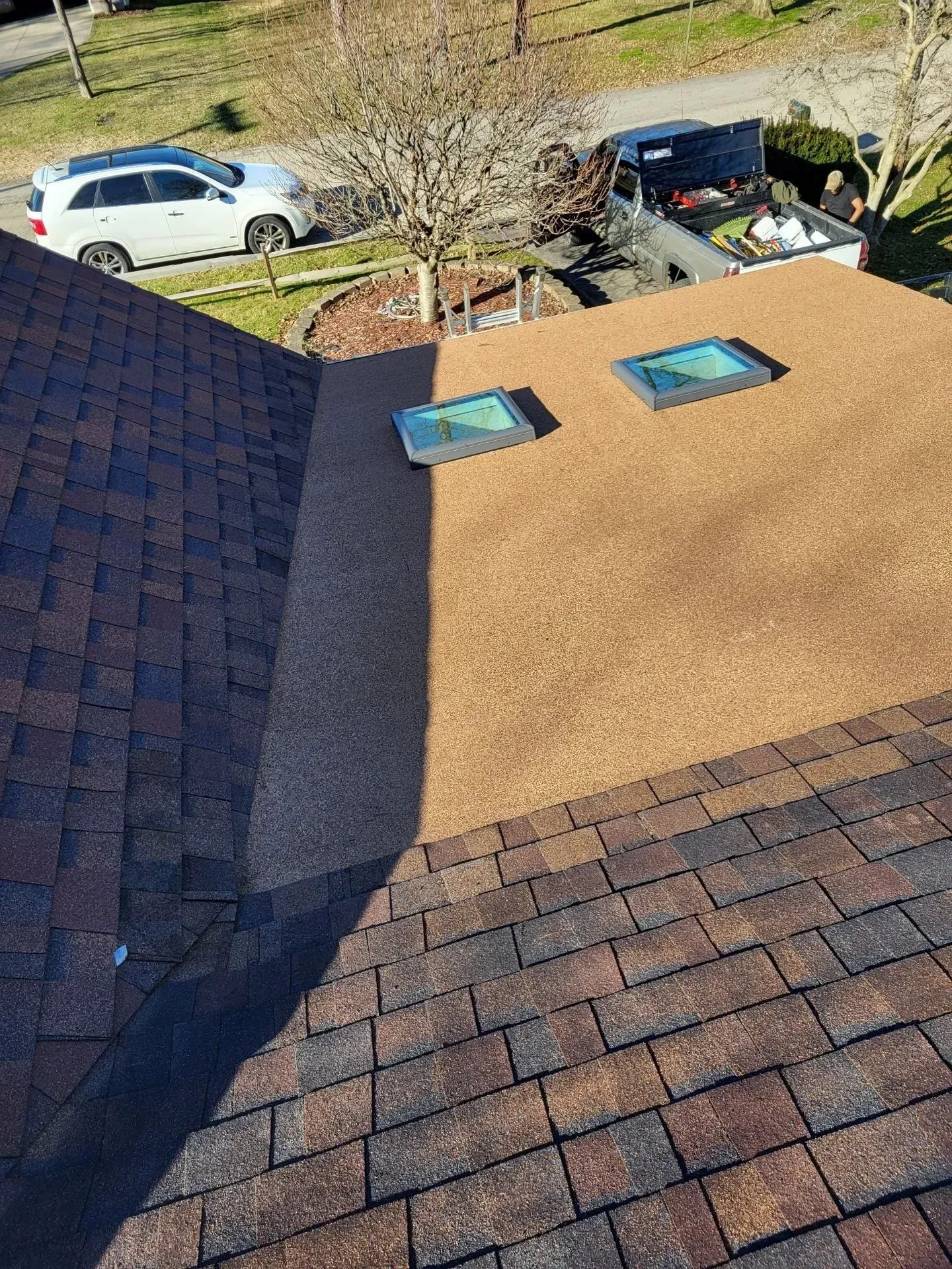Overhead view of a roof with brown and tan shingles, two skylights, and a vehicle in the background.