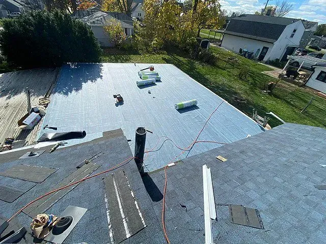 Roofing work in progress on a residential house; blue underlayment and shingles are visible.