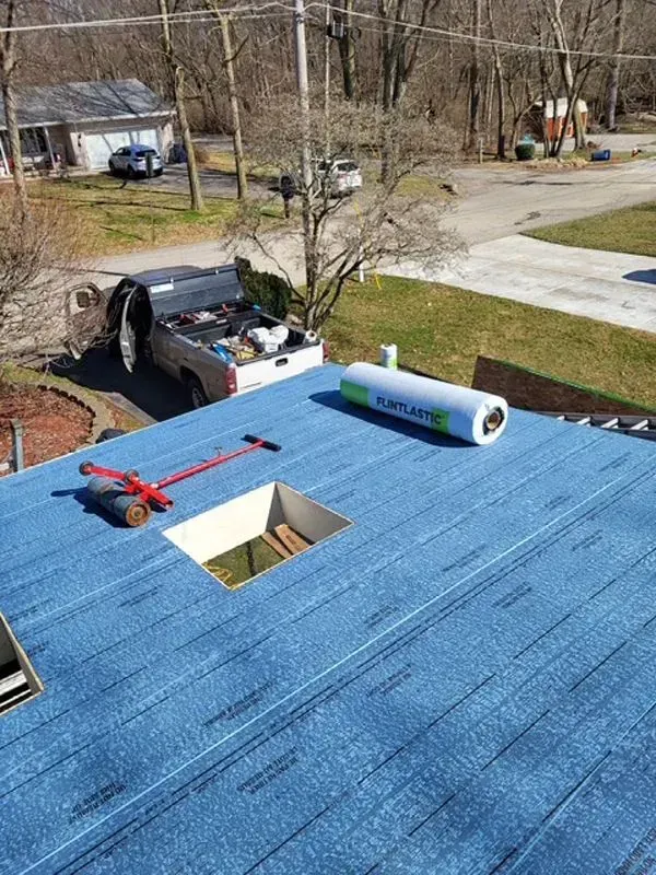 Rooftop with blue underlayment, open skylight, and rolled roofing material; truck in the background.