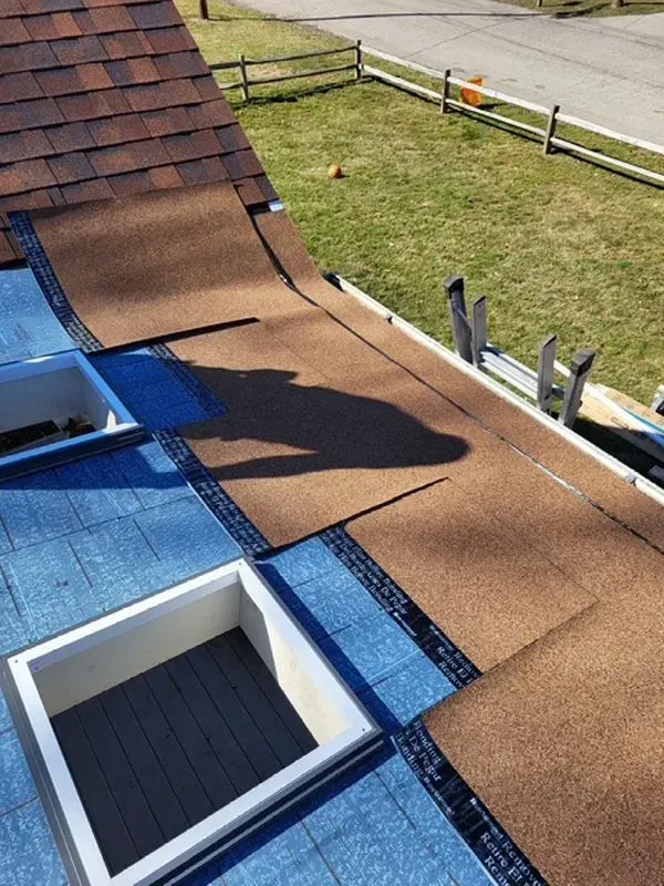 Roof being repaired, showing brown shingles being laid over blue underlayment near a skylight.