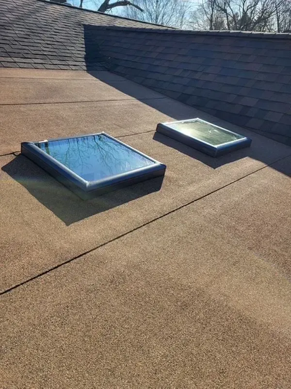 Two rectangular skylights on a flat, brown roof with a dark shingle roof in the background.