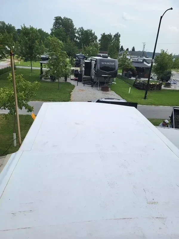 White RV roof with a campground scene in the background; RVs, trees, and cloudy sky.