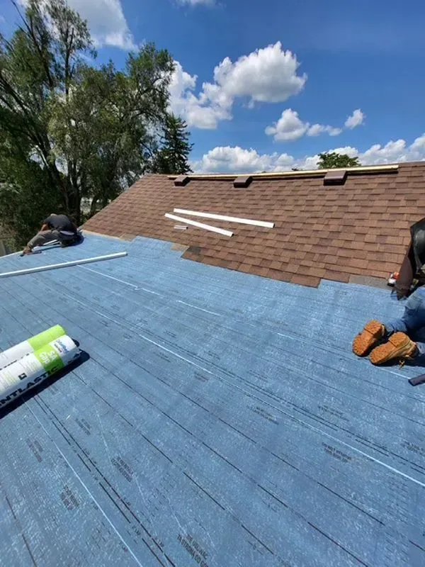 Roofers installing shingles on a roof. Blue underlayment visible, brown shingles being laid. Sunny day.