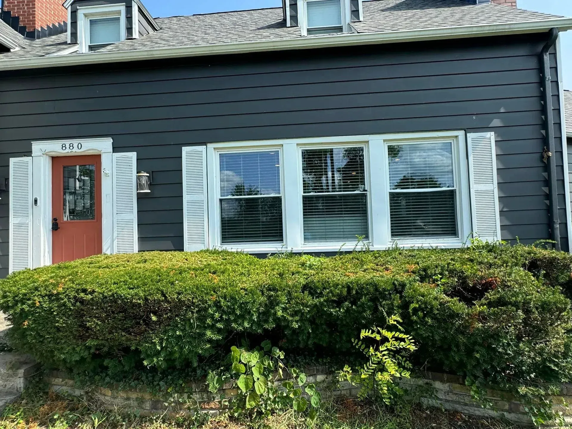 Dark-gray house with white trim, shutters, and windows; a red door. A green hedge in front.