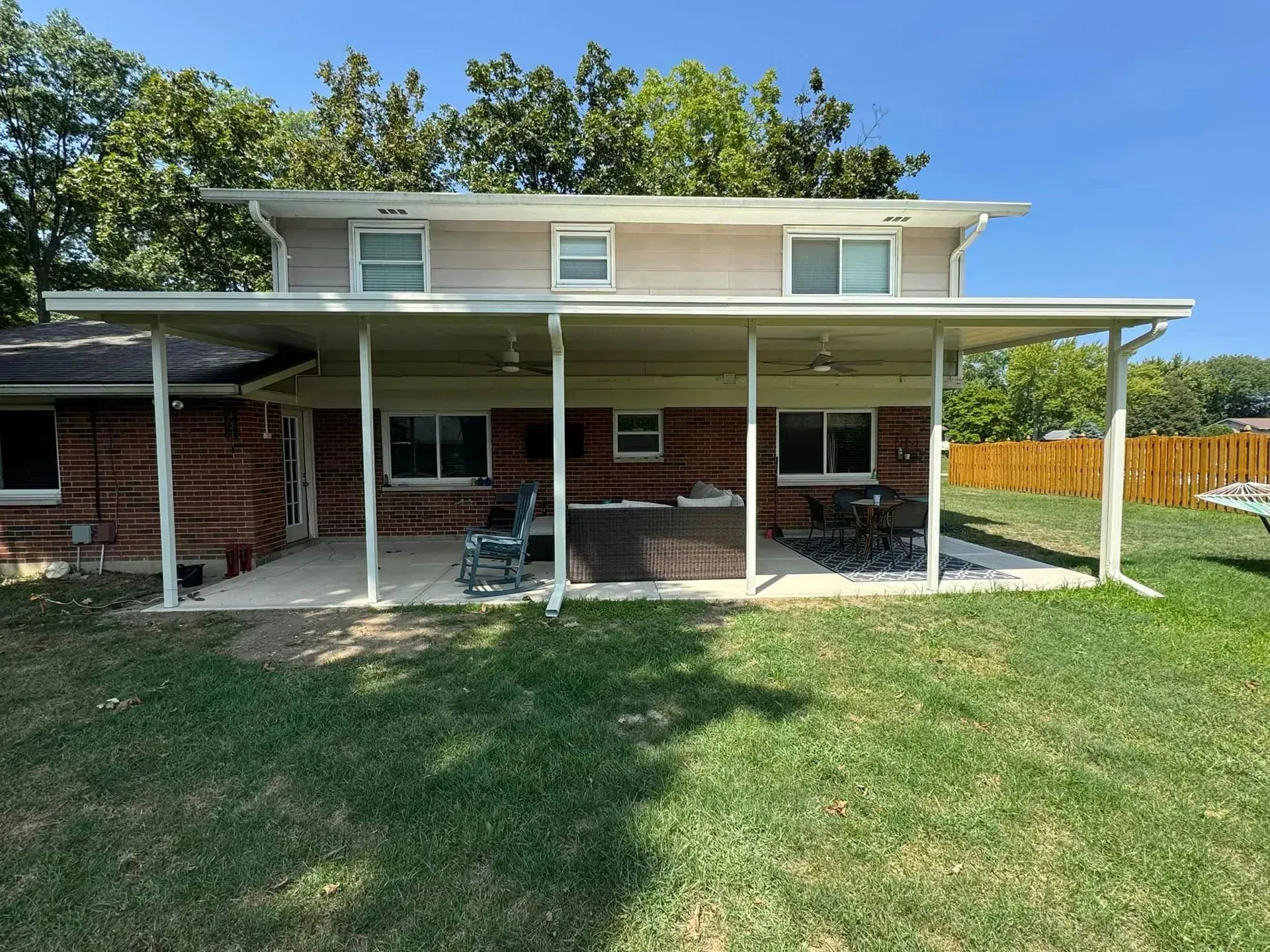 A two-story home with a patio cover over a concrete patio; red brick exterior, green grass, and trees in the background.