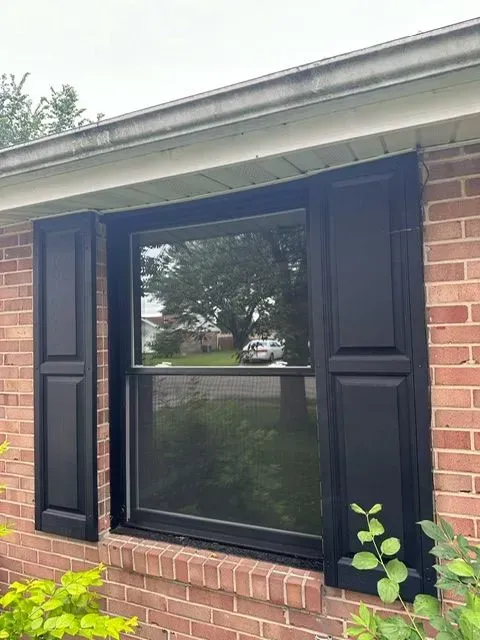 Window with black shutters on a brick building, view of a yard and trees through the glass.