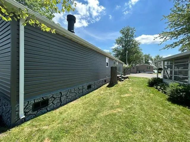 Gray-sided mobile home with a black chimney and stone-like skirting on a grassy lawn under a blue sky.