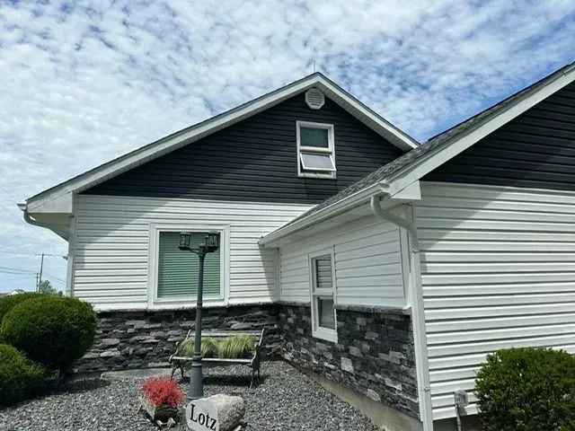 Two-story house with black and white siding, stone base, and green bushes against a cloudy sky.