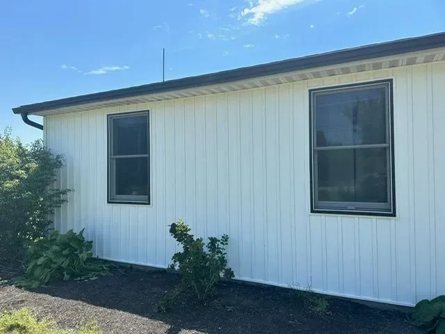 White-sided building with two windows and dark trim under a blue sky, shrubs in front.