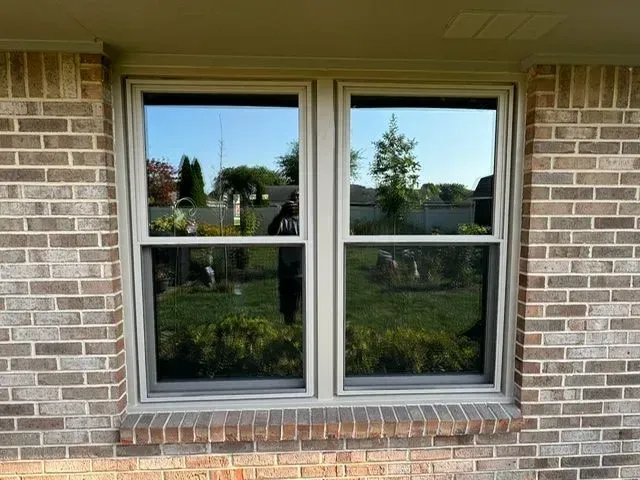 Double-hung windows on brick house reflecting the backyard; green grass, trees and a blue sky are visible.