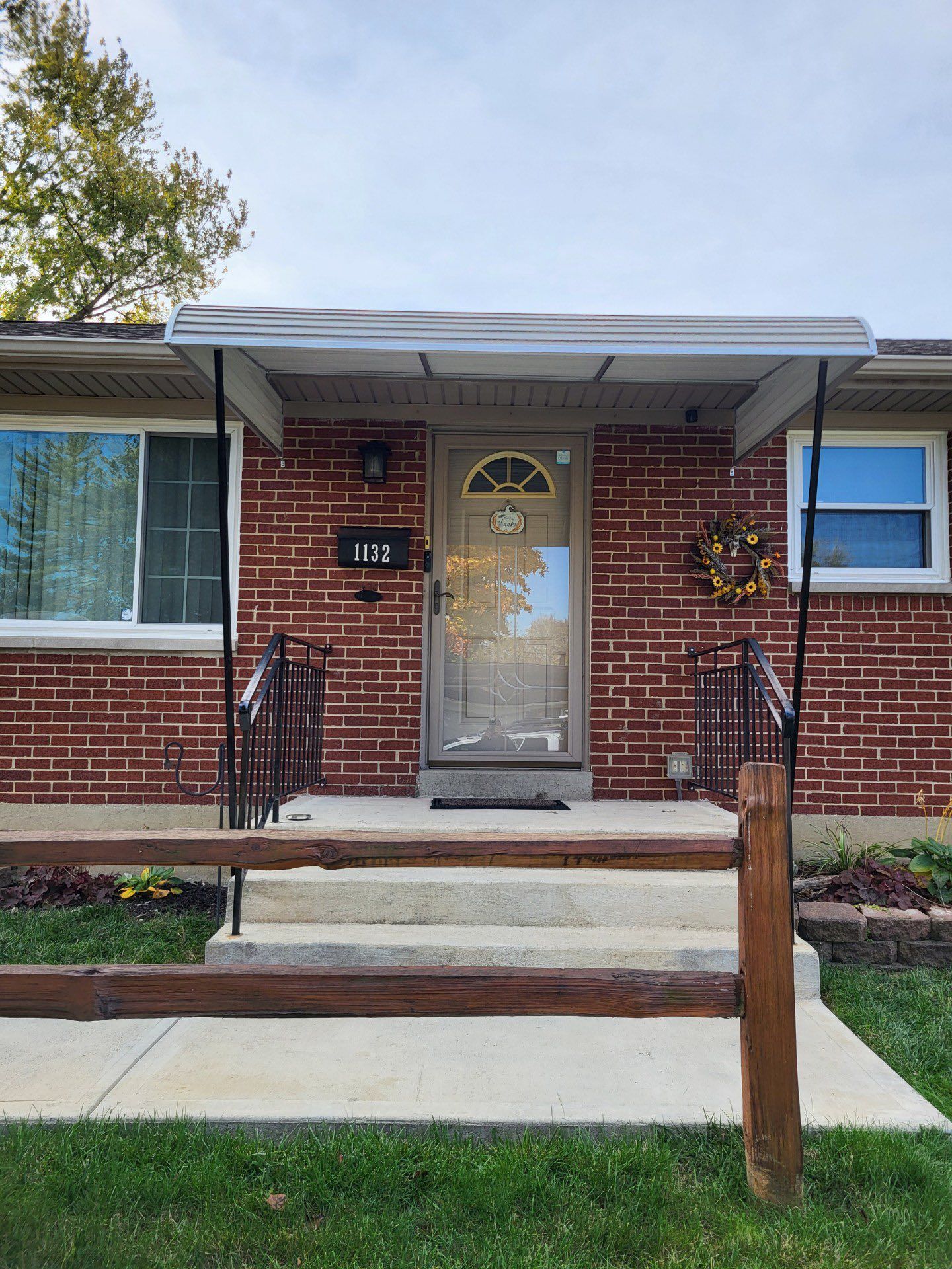 Brick house with a front porch, door, windows, and small awning. Wooden railing and concrete walkway.
