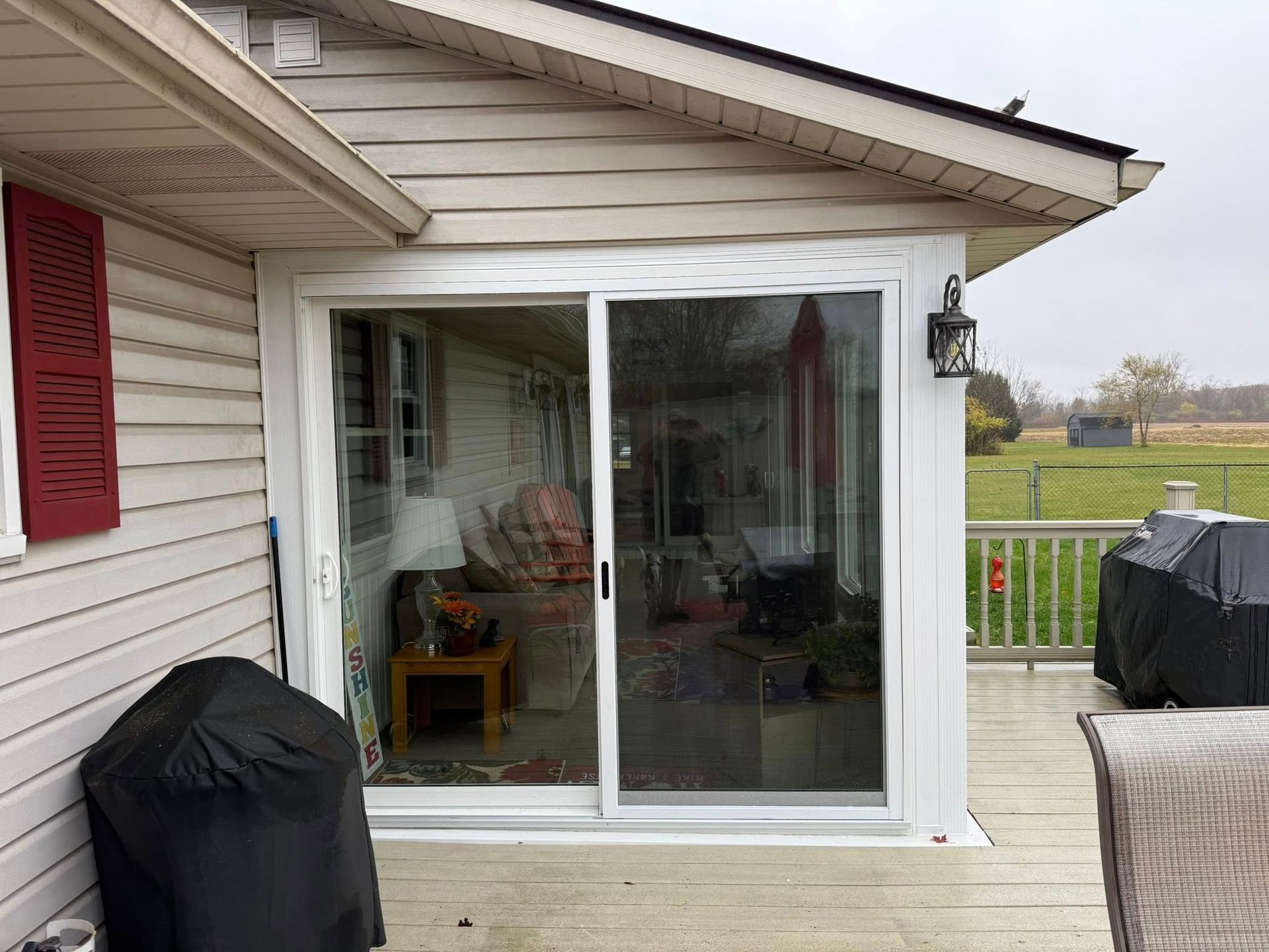 White sliding glass door on a deck, leading into a room with furniture. Red shutter and porch.