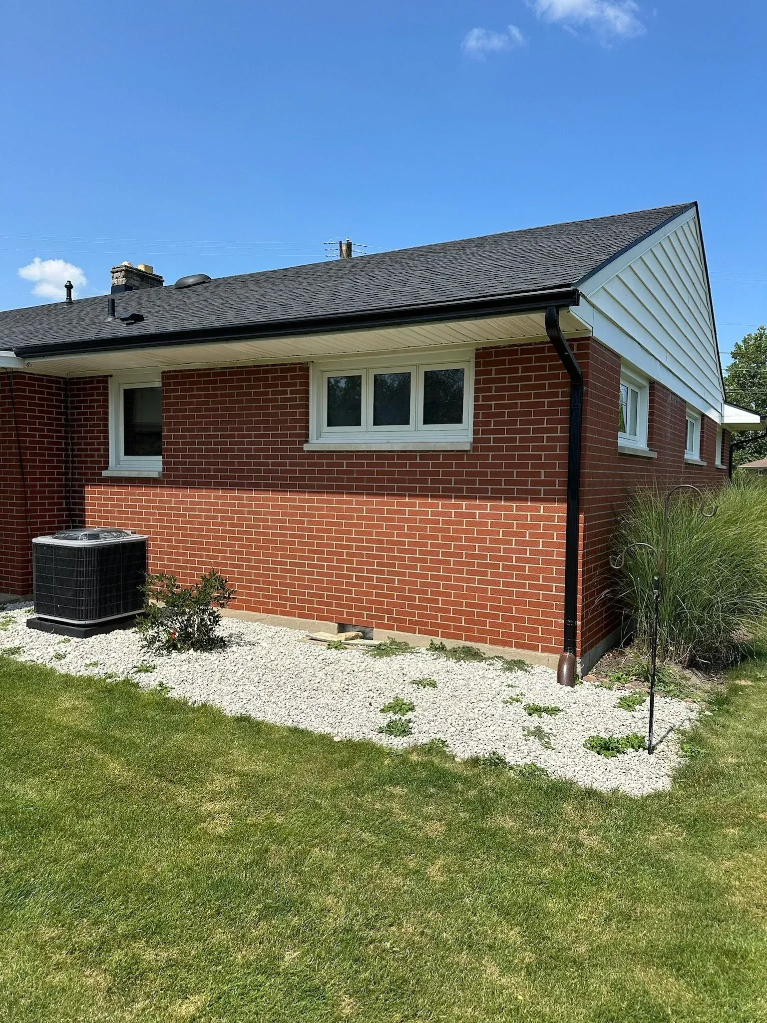 Red brick house with black roof, white trim, and AC unit on grass with rock border.