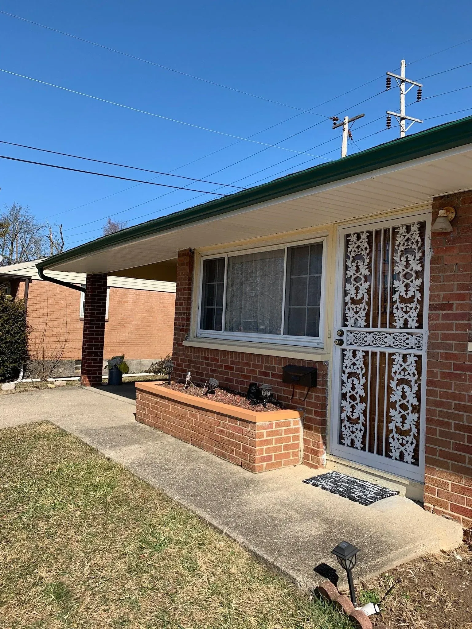 Brick house with a covered porch, security door, and small planter box. Concrete pathway leads to the entrance.