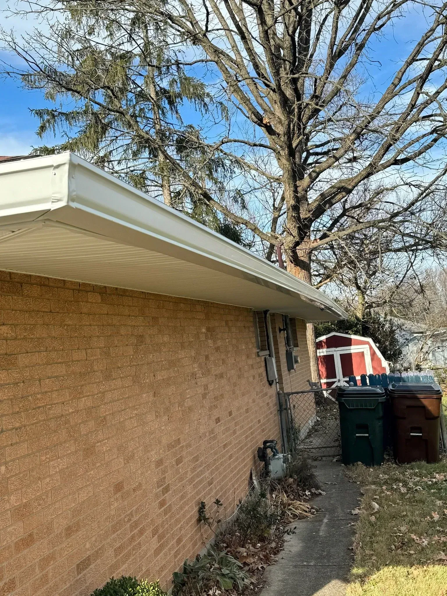 Brick building exterior with white trim, gutters, and a tree against a blue sky. Trash cans are visible.