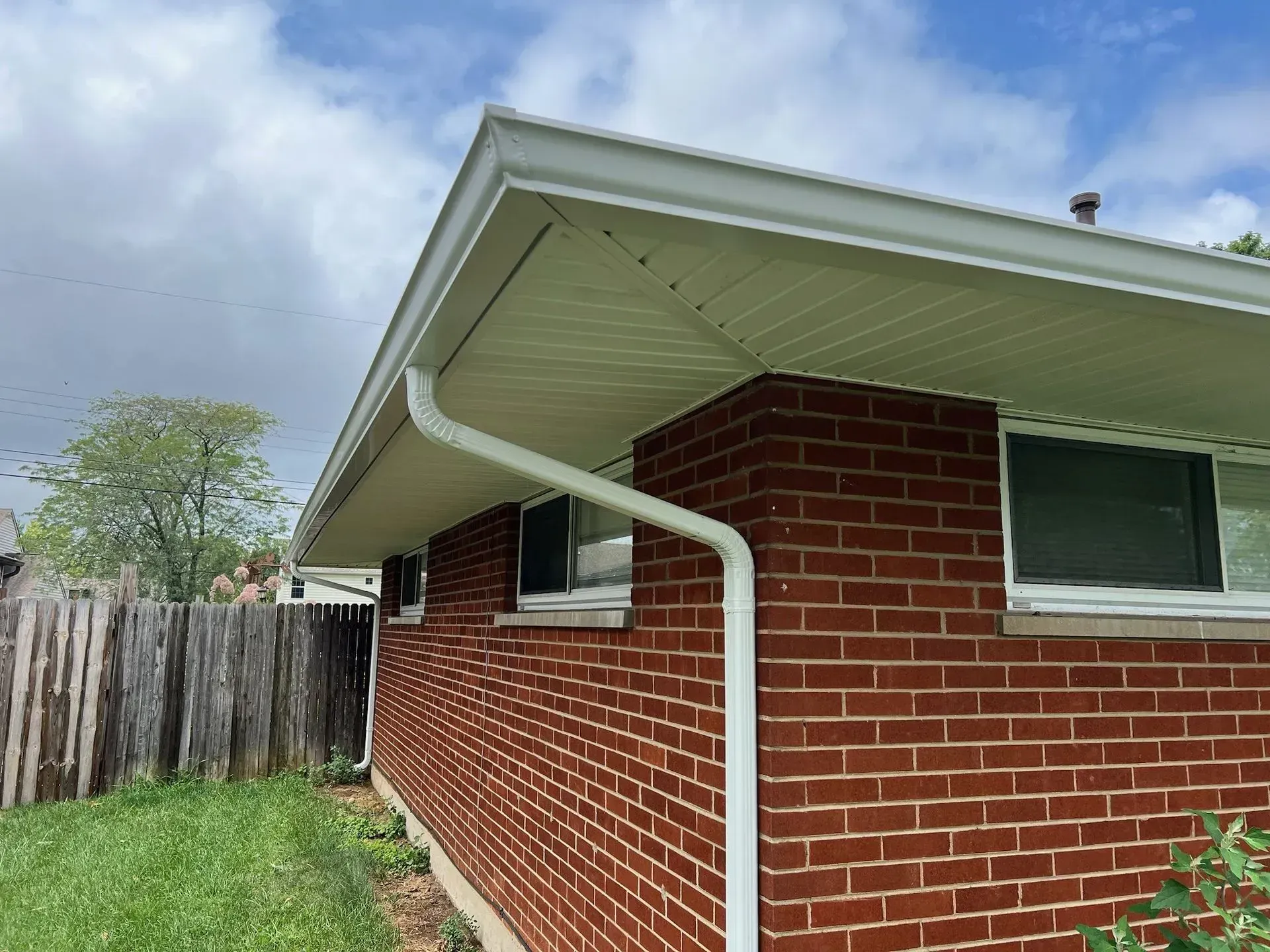 Red brick house with white gutters and trim, green lawn, and a wooden fence under a cloudy sky.