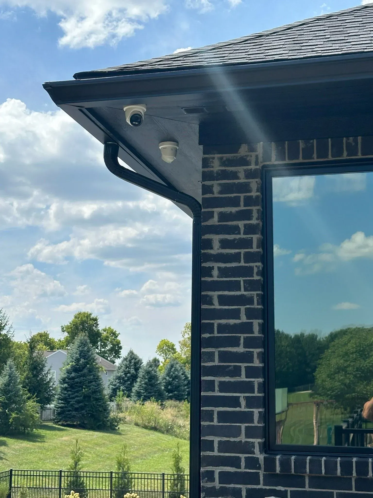 Two security cameras mounted on a dark brick building's corner under a black gutter and roof, with a window reflecting the sky.