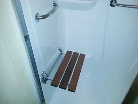 Shower with grab bars and fold-down wooden seat. White walls, chrome fixtures.