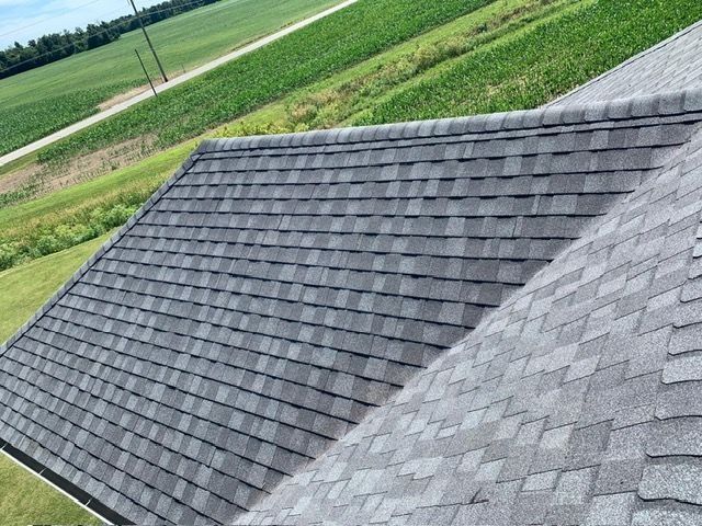 Gray asphalt shingle roof on a house, with a green field in the background.