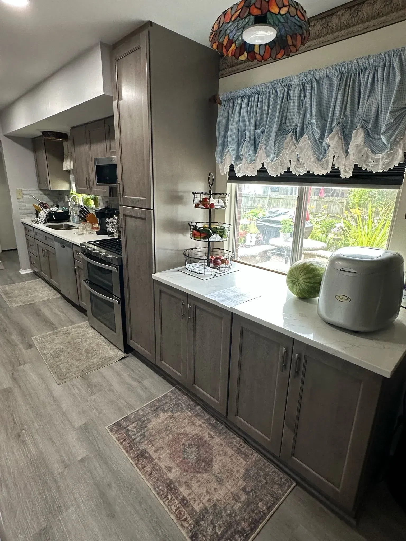 Kitchen with gray cabinets, window with lace curtain, countertop, rug, and appliances.