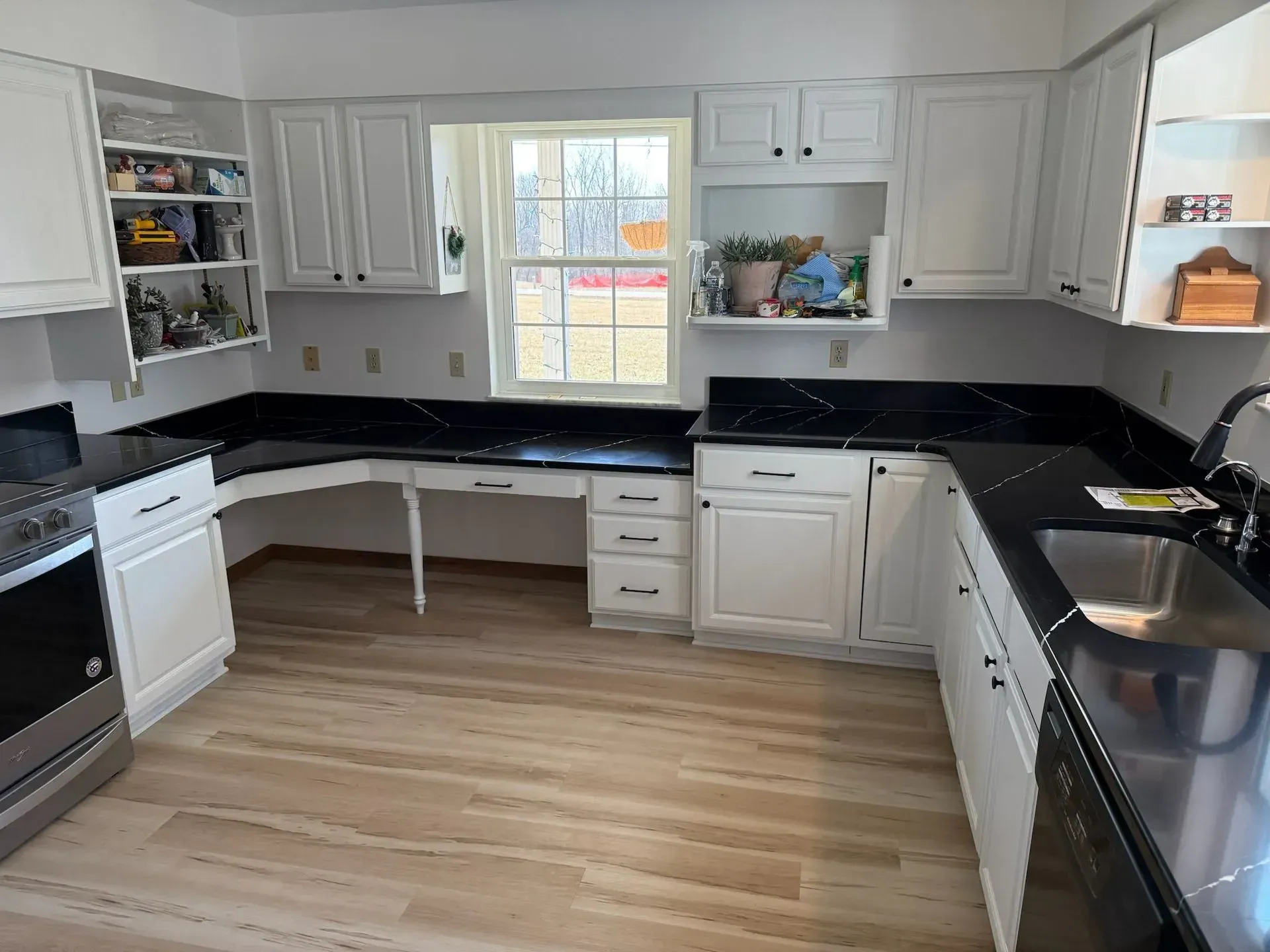 White kitchen with black countertops, accessible desk area by window, light wood floors.