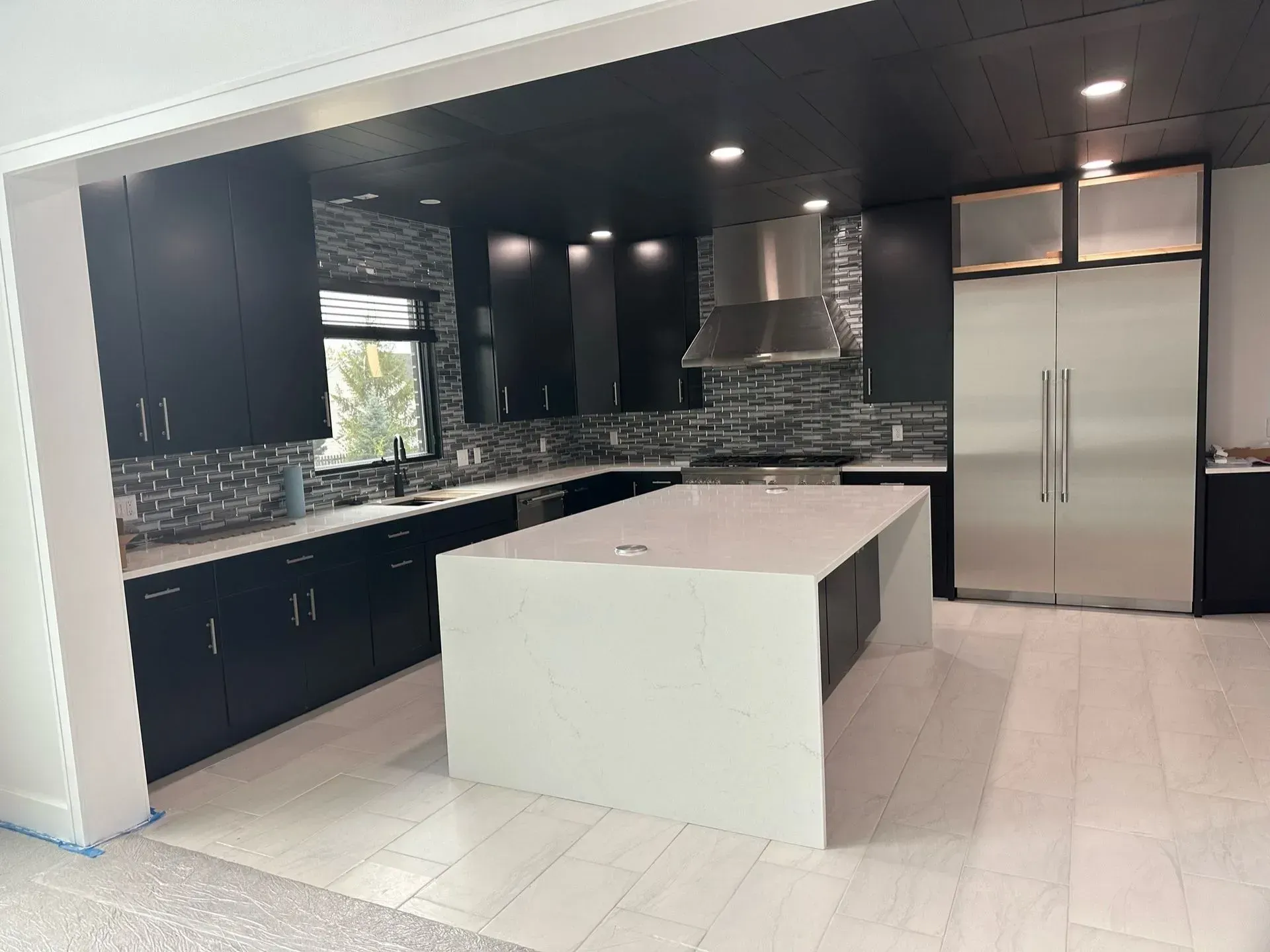 Modern kitchen with black cabinets, white island, stainless steel refrigerator, and tiled backsplash.