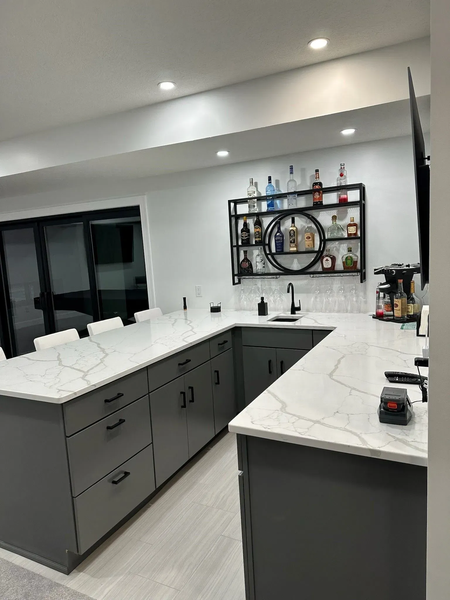 Gray kitchen cabinets with white countertops, a bar area with liquor bottles, and modern lighting.