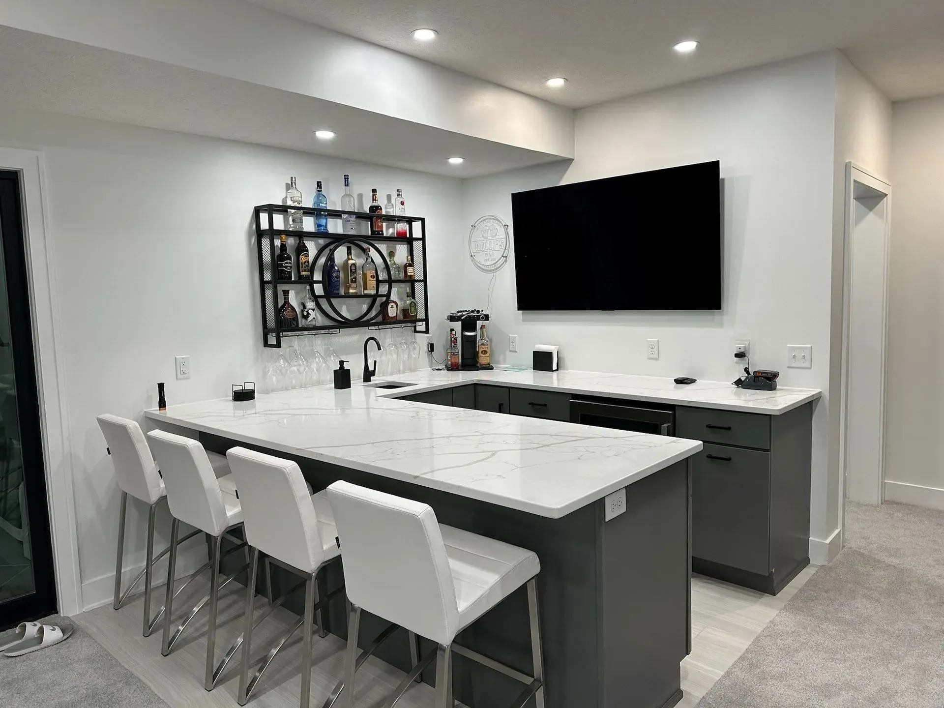 A modern home bar with white countertop, gray cabinets, bar stools, liquor, and a mounted TV.