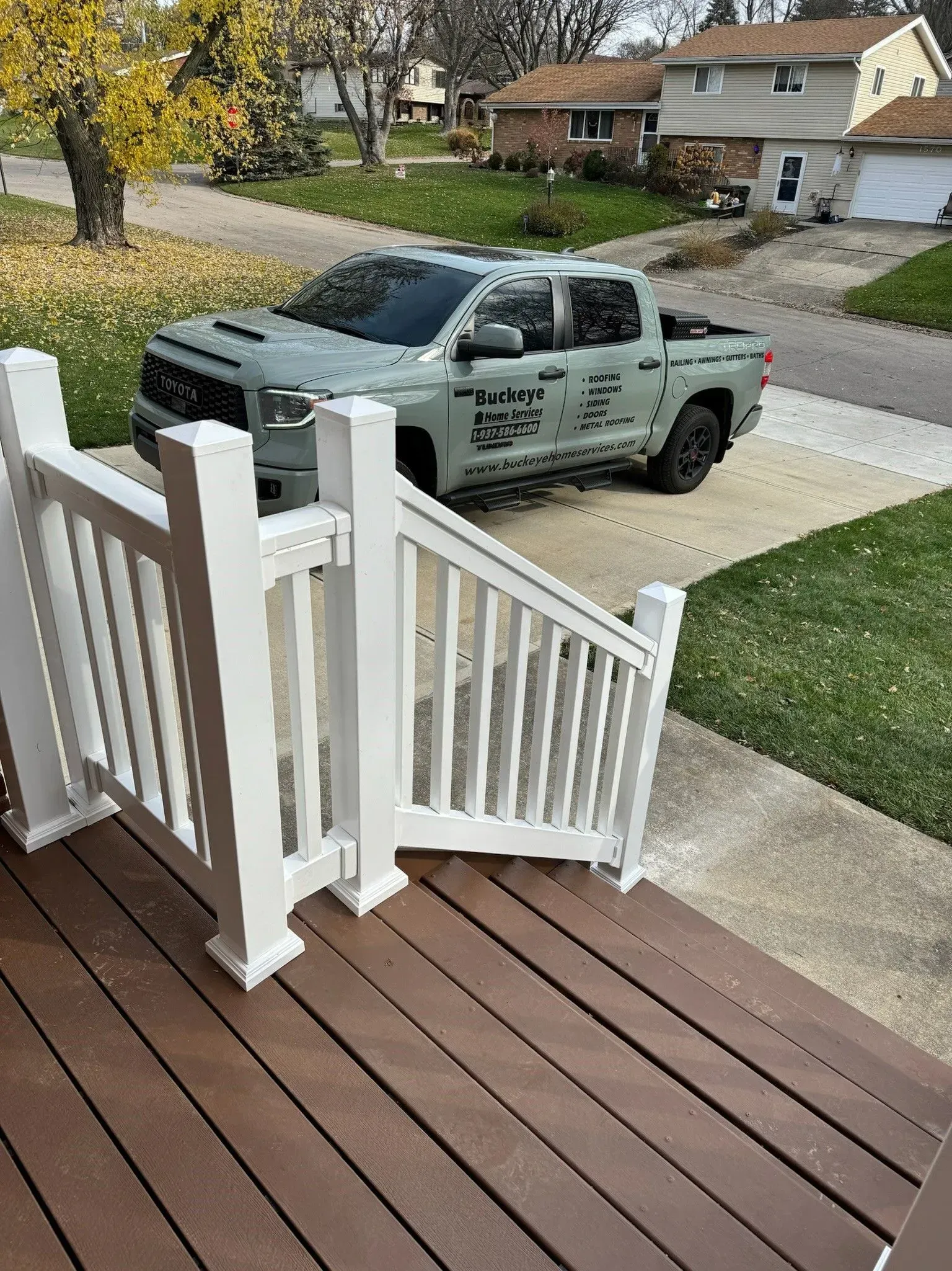White railing on a wooden porch leads to a green truck parked on a driveway in front of houses.