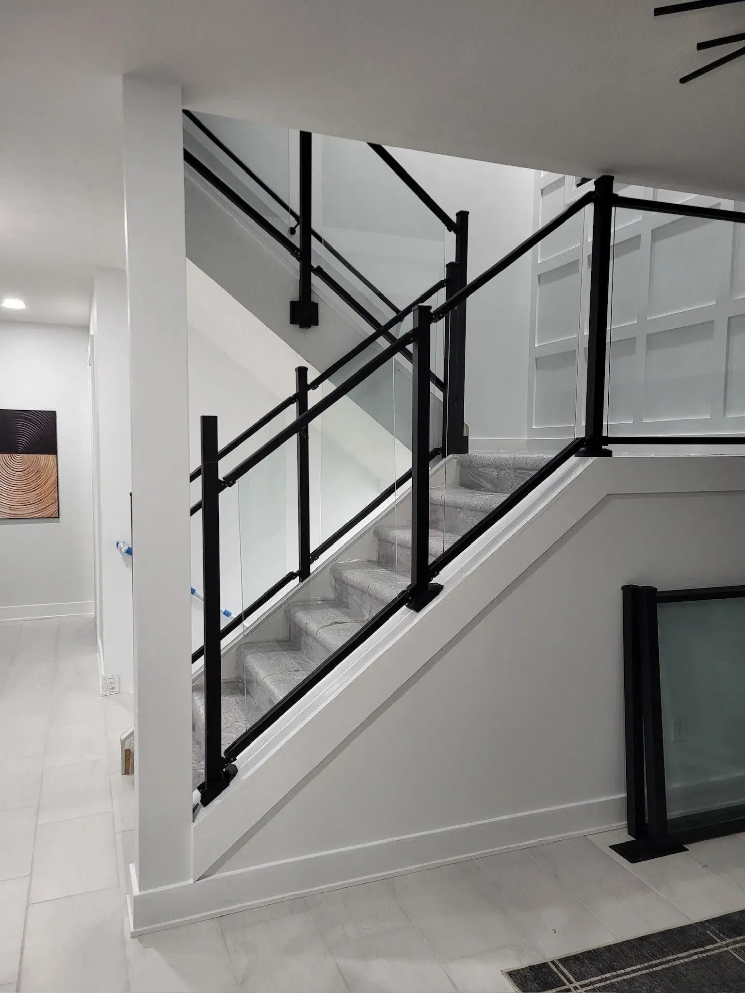 Staircase with black metal railing and glass panels. Gray carpeted steps, white walls, and a minimalist design.