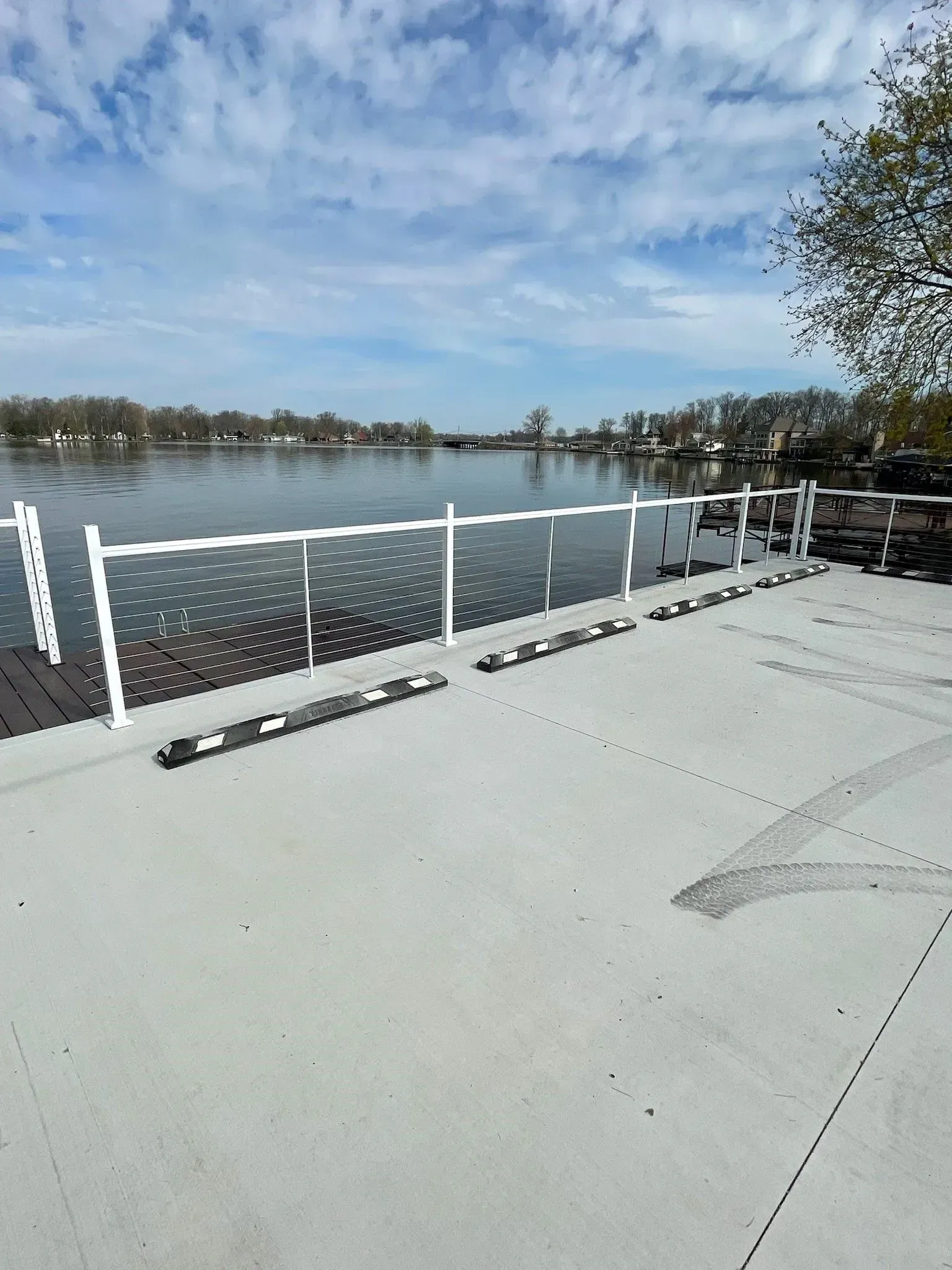 Concrete pier with white railing overlooks calm water and cloudy sky.