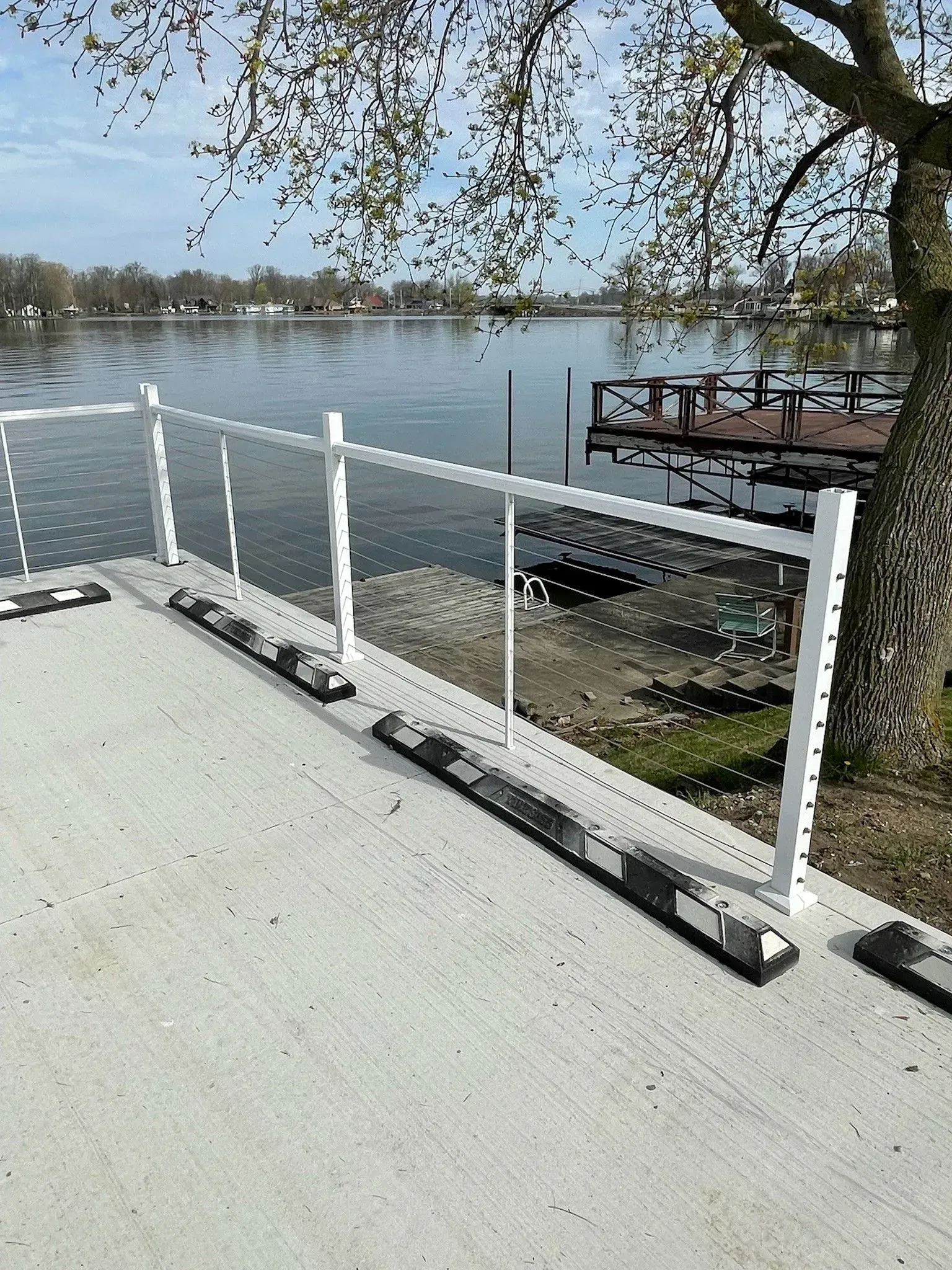 White railing on a concrete walkway bordering a lake with a wooden dock and trees.