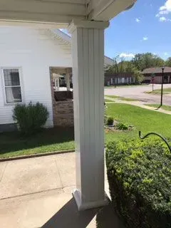 White porch column next to a green lawn with a building and street in the background. Sunny day.