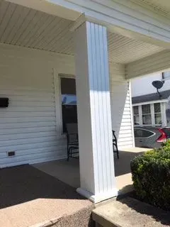White porch with a fluted column supporting the roof, next to a door and bush.
