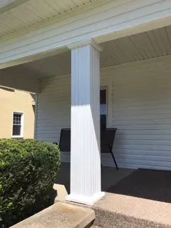 White porch with a column, a bush, and a light-colored building in the background.
