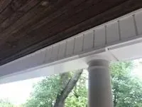 White porch column supporting a white soffit with vertical siding, beneath a brown wooden ceiling.