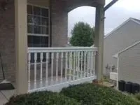 White railing on a porch with tan columns and a window. Green bush in the background.