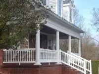 White porch with columns, railing, and stairs on a brick-faced house, tree in front, blue sky.