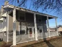 Two-story gray house with white porch and railing. Sunny day, bare tree branches above.
